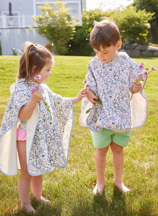 Two young children standing barefoot on grass, wearing matching floral hooded ponchos and holding purple flowers on a sunny day.