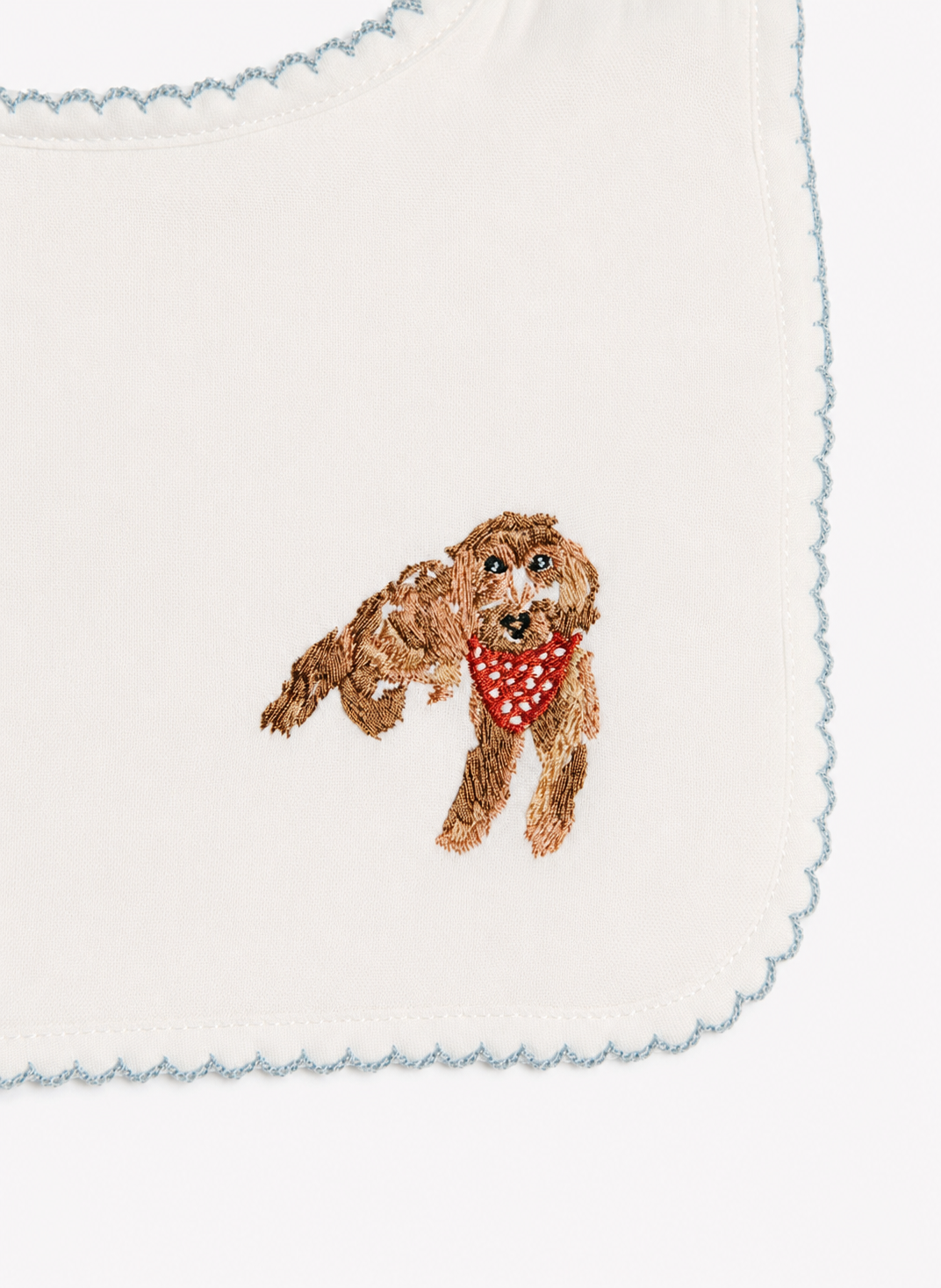 Close-up of a white baby bib with scalloped edges and an embroidered brown dog wearing a red polka-dot bandana.