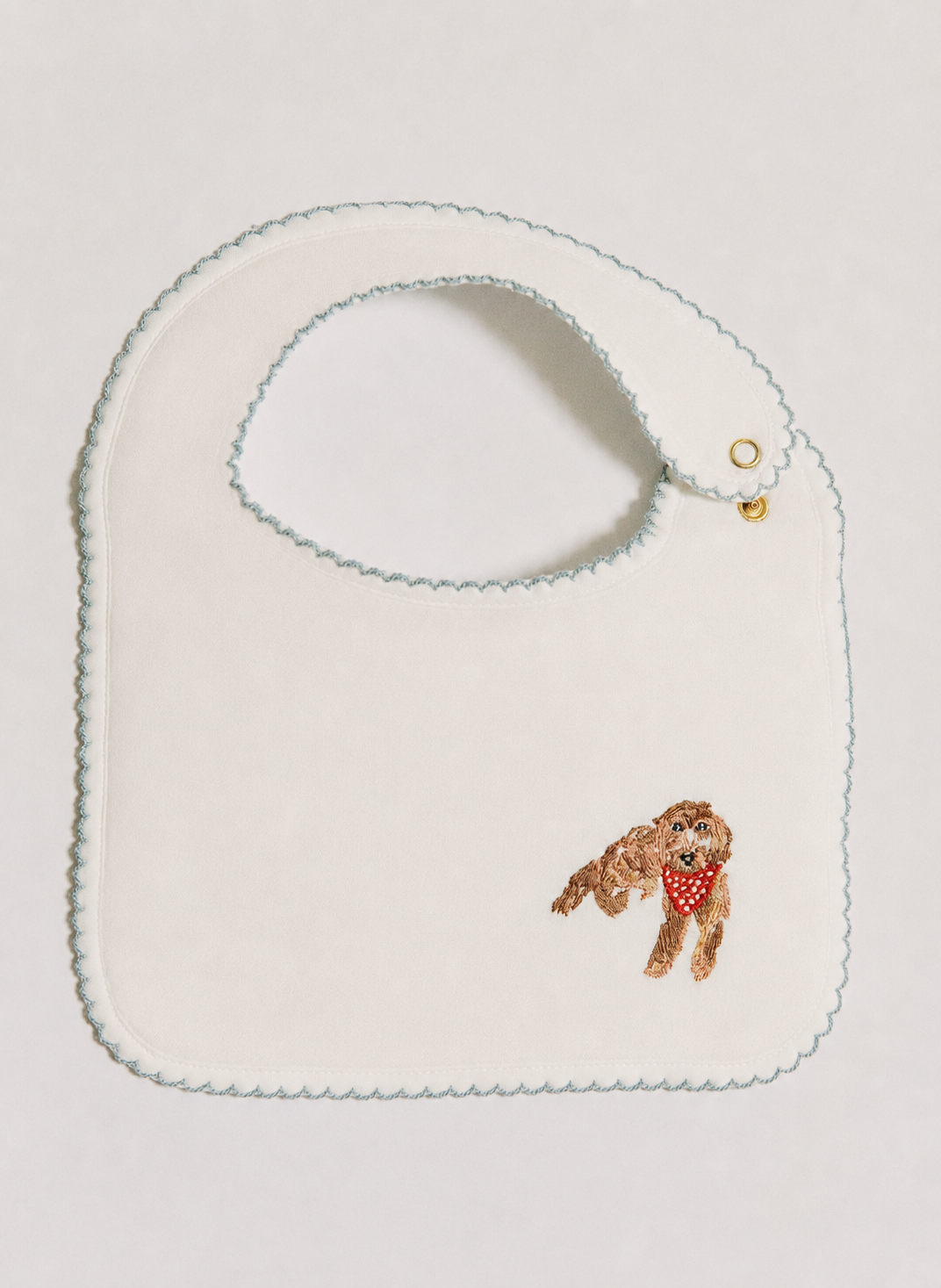 An off-white baby bib with a scalloped edge, featuring a small embroidered brown dog wearing a red bandana in the bottom right corner.
