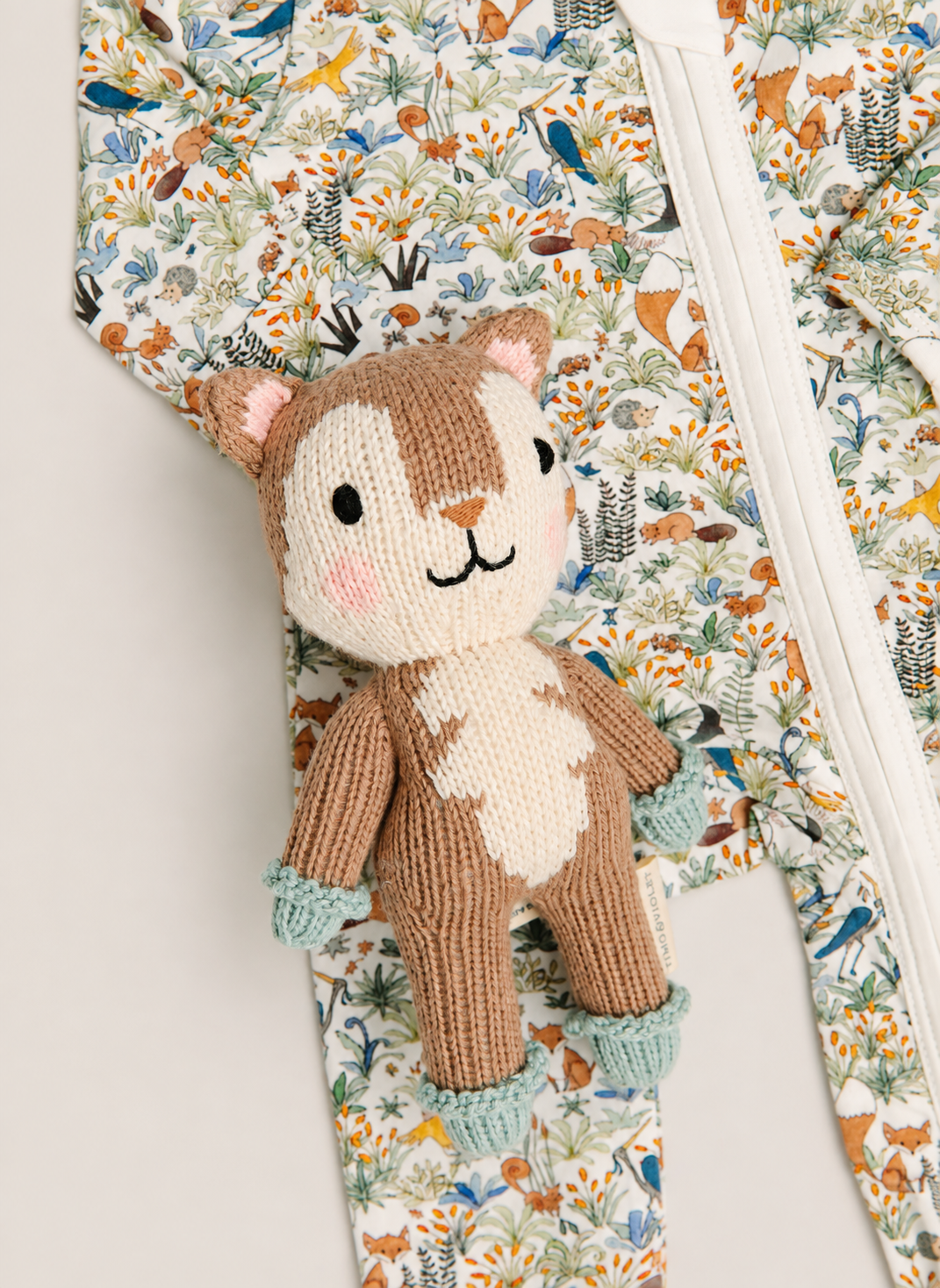A close-up of a knitted deer plush toy placed on top of a floral and woodland print zippered garment. The toy is brown and cream with pink cheeks and light blue booties, while the background fabric has a multicolor nature print with birds, foxes, and foliage.