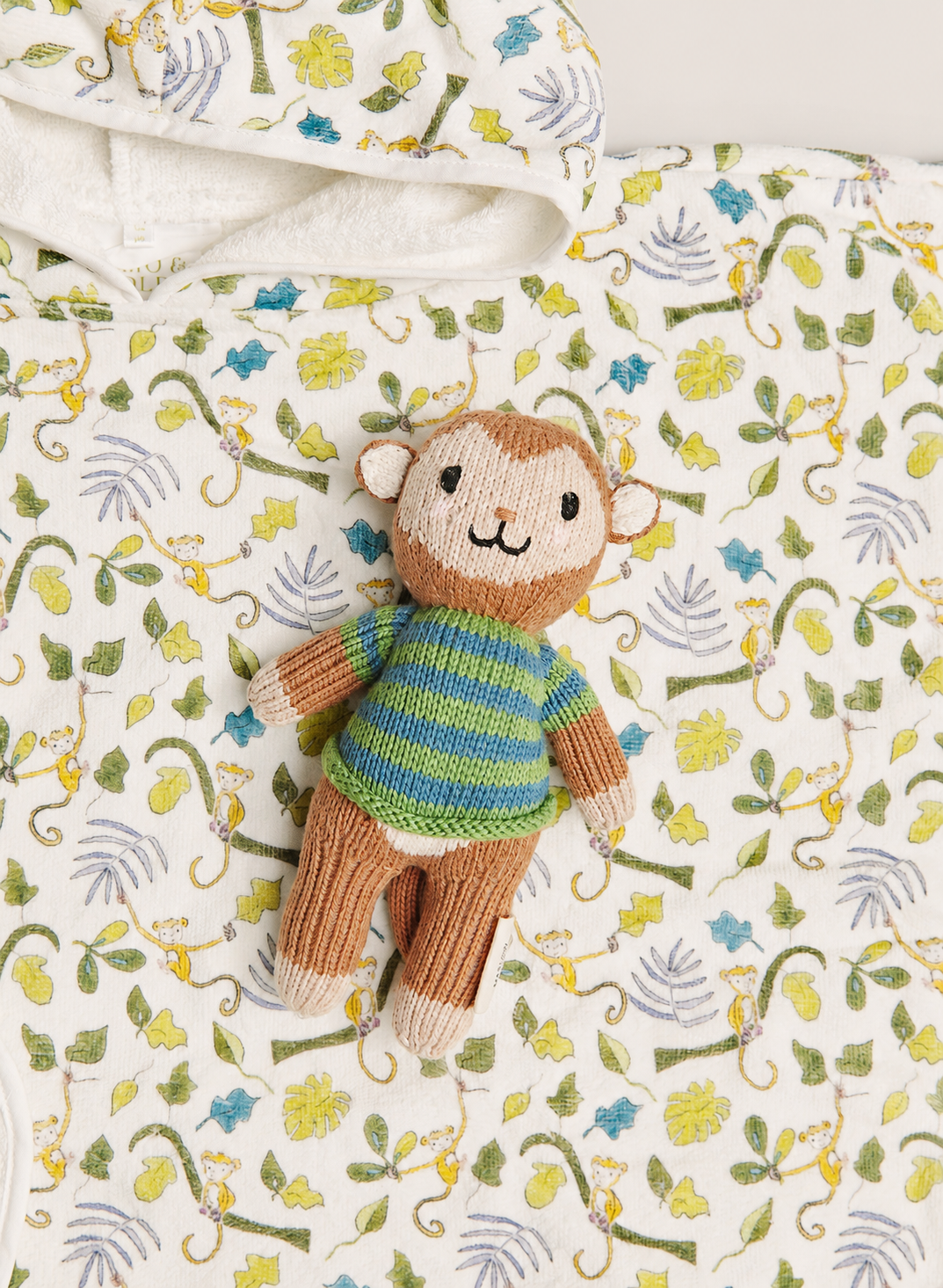 A small knitted monkey toy wearing a striped green and blue sweater, placed on a white quilted blanket with a yellow and green jungle leaf pattern.