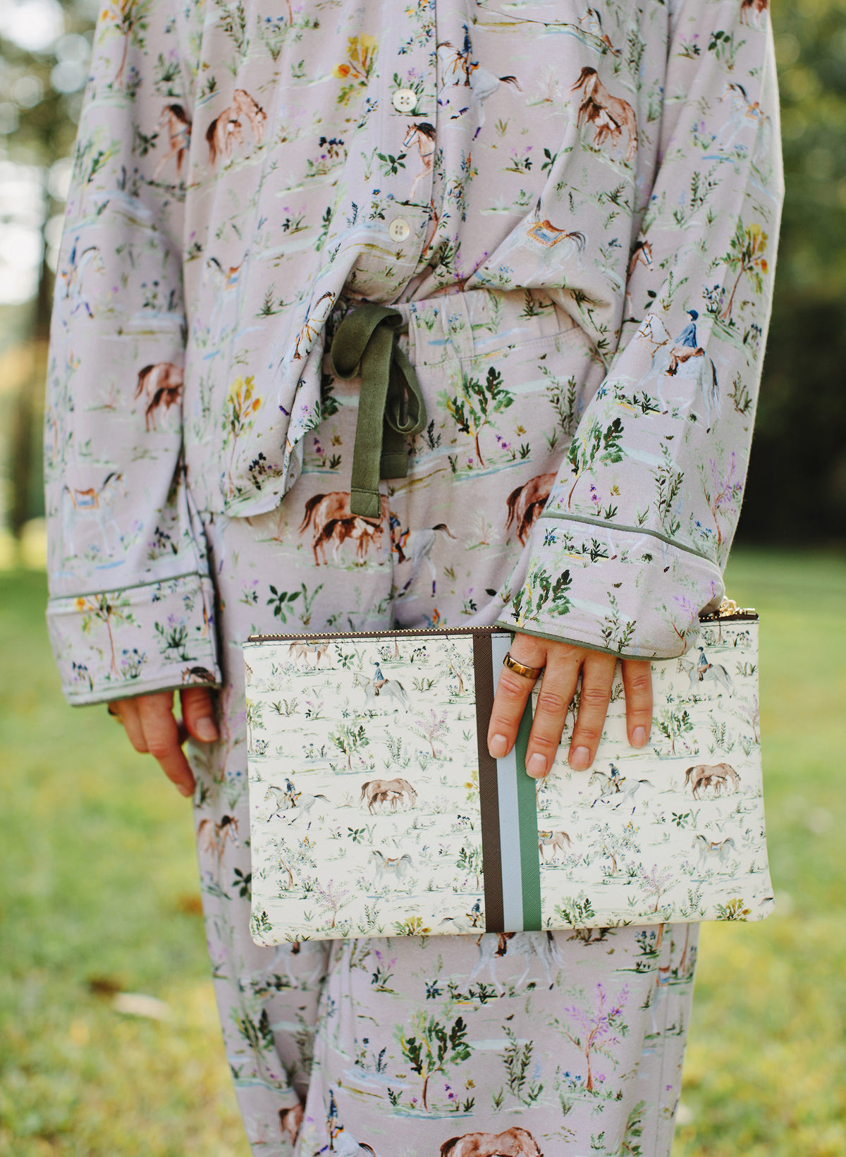 A person wearing a matching pajama set with an all-over horse and floral print, holding a matching clutch bag outdoors on a grassy lawn.