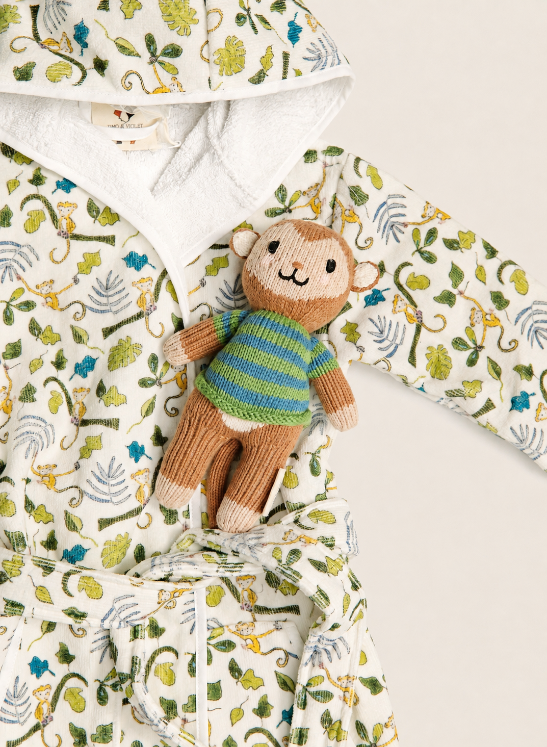 A baby hooded robe with a jungle-themed print in green, yellow, and blue on an off-white background, styled with a small plush monkey toy wearing a striped sweater.