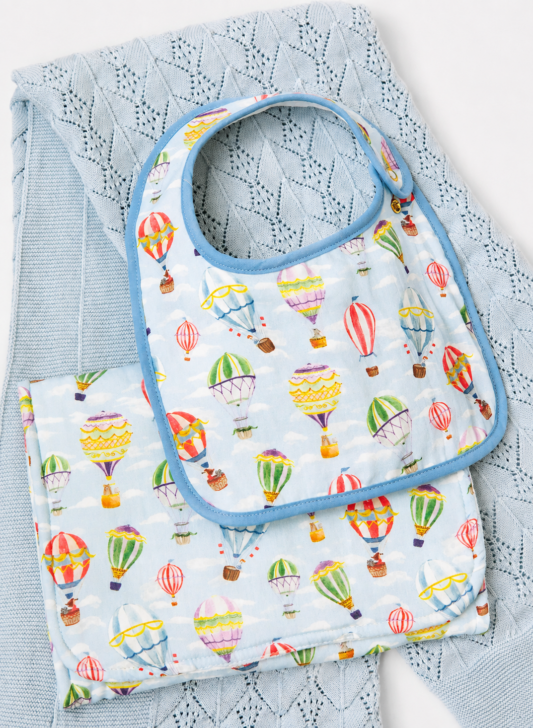 A baby bib and matching square cloth with a colorful hot air balloon print on a pale blue background, styled on top of a light blue textured blanket.