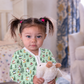 A young child with brown pigtails wearing a light green pajama shirt with a woodland animal print, holding a white plush toy in a softly lit bedroom.