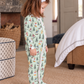 A young child wearing a light green footed pajama with a festive Christmas print of trees, Santa Claus, and snowmen, standing in a cozy bedroom setting.
