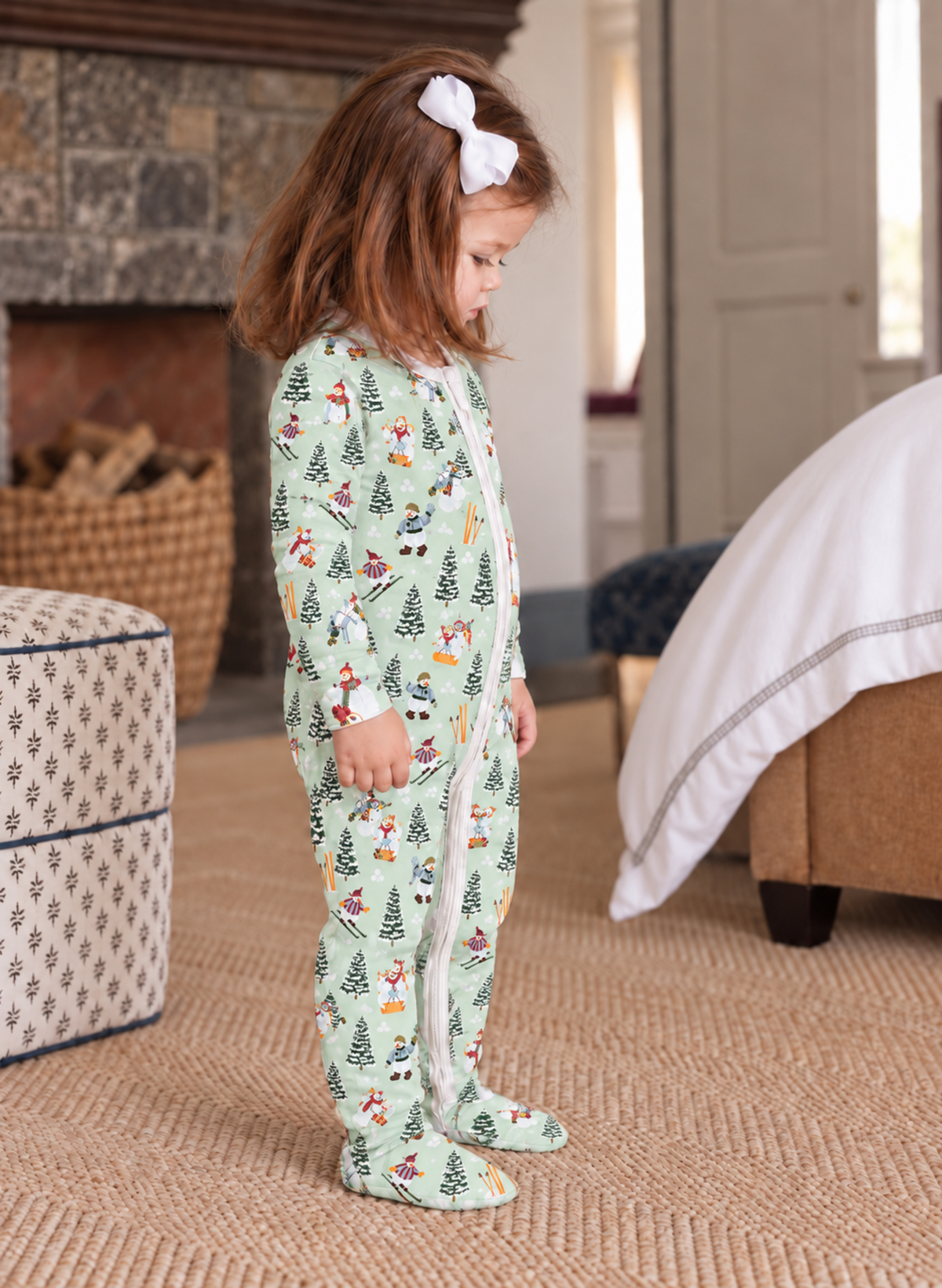A young child wearing a light green footed pajama with a festive Christmas print of trees, Santa Claus, and snowmen, standing in a cozy bedroom setting.