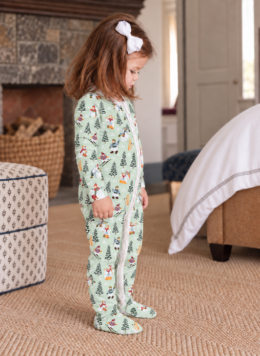 A young child wearing a light green footed pajama with a festive Christmas print of trees, Santa Claus, and snowmen, standing in a cozy bedroom setting.