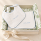 A baby gift set featuring a white bib and matching square cloth with light blue scalloped edges and embroidered 'amore' text, presented in a cream gift box with green patterned tissue and a cream ribbon.
