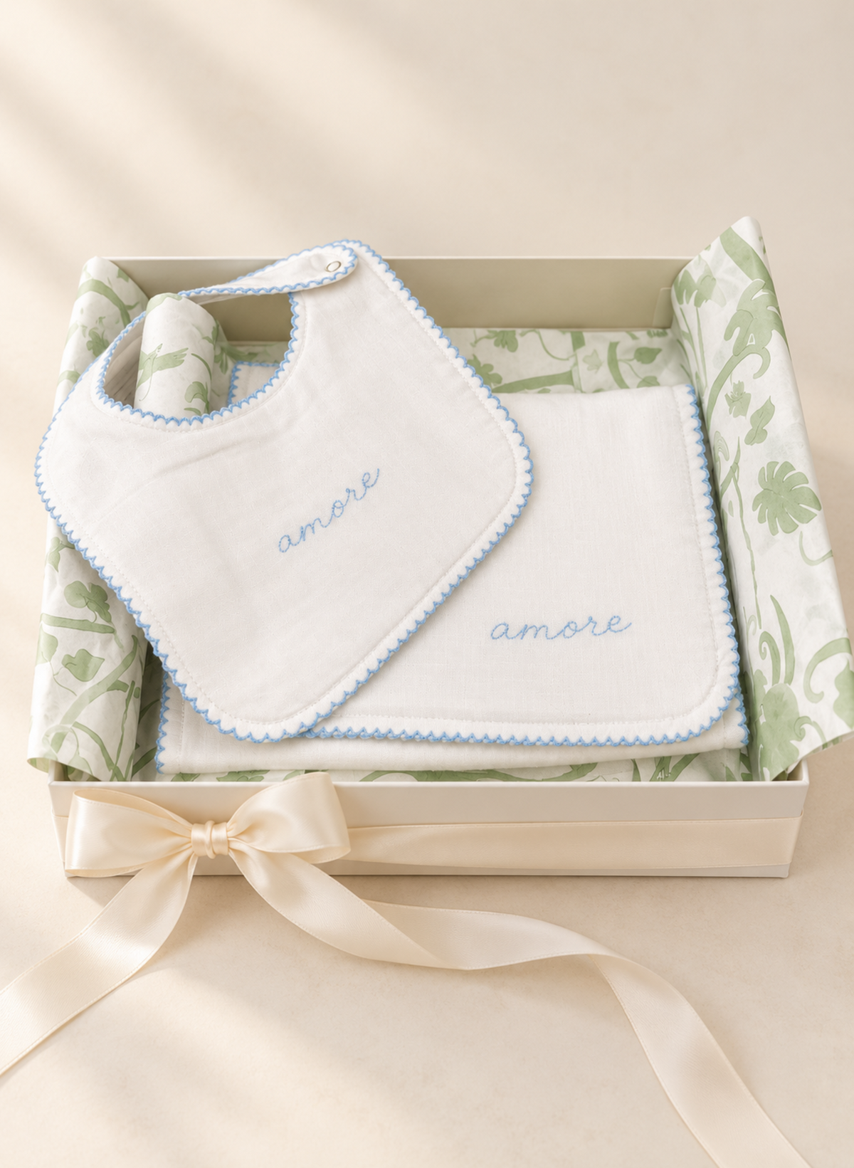 A baby gift set featuring a white bib and matching square cloth with light blue scalloped edges and embroidered 'amore' text, presented in a cream gift box with green patterned tissue and a cream ribbon.