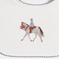 A close-up of a white fabric product, likely a bib or scarf, featuring a central embroidered design of a white horse with brown details and a rider in light blue and white. The fabric has a scalloped gray border.