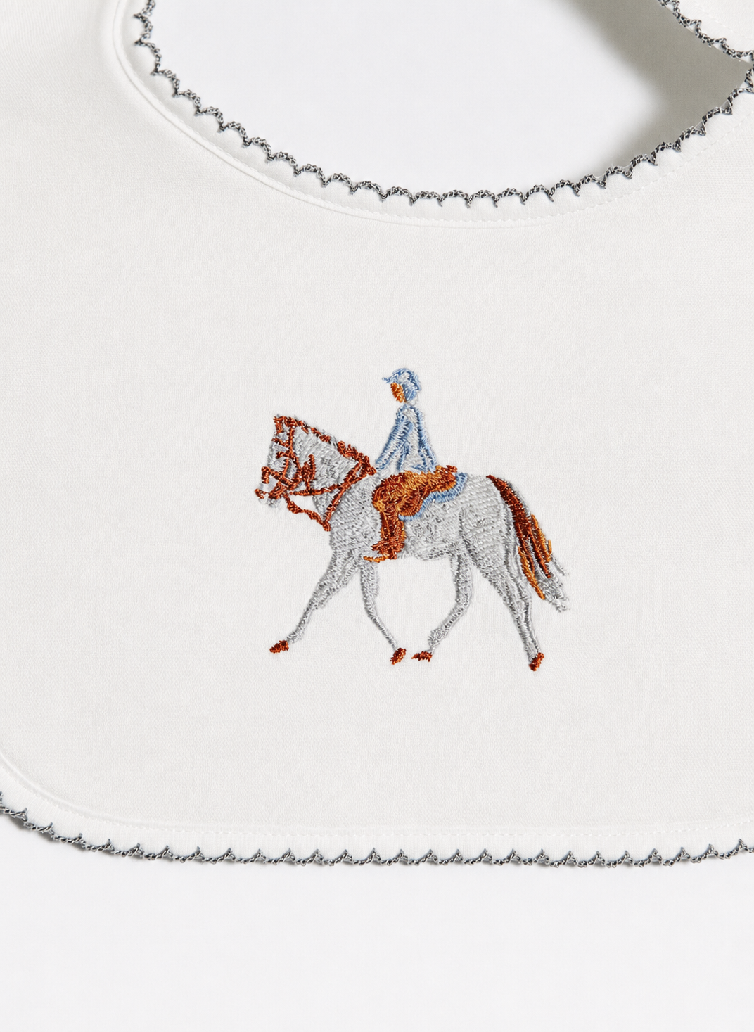 A close-up of a white fabric product, likely a bib or scarf, featuring a central embroidered design of a white horse with brown details and a rider in light blue and white. The fabric has a scalloped gray border.
