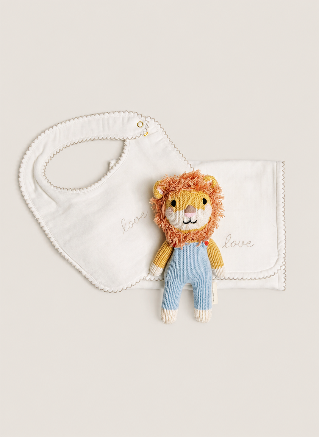 A baby gift set featuring a white embroidered bib with scalloped edges, a matching white muslin baby blanket with 'love love' embroidered in gold, and a small plush lion toy wearing yellow and blue knitted overalls, arranged on a light beige background.