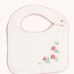 An off-white cotton baby bib with a scalloped pink border, featuring three embroidered red poppies with green leaves on the lower right and a gold snap button on the neck strap.