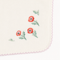 Close-up of a white or off-white fabric accessory with scalloped pink stitching, embroidered red poppy flowers with green stems, and a small gold metal ring at the top corner.
