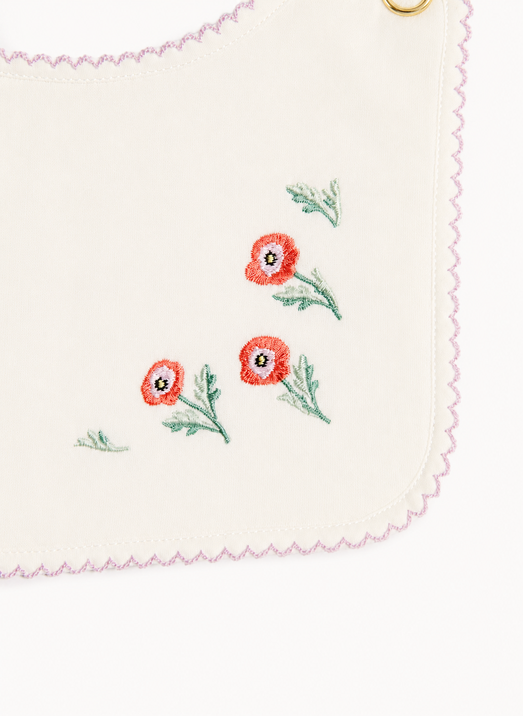 Close-up of a white or off-white fabric accessory with scalloped pink stitching, embroidered red poppy flowers with green stems, and a small gold metal ring at the top corner.
