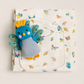 A folded white muslin baby blanket with a colorful butterfly and peacock pattern, featuring a front pocket with a white drawstring and a blue knitted peacock plush toy tucked inside.
