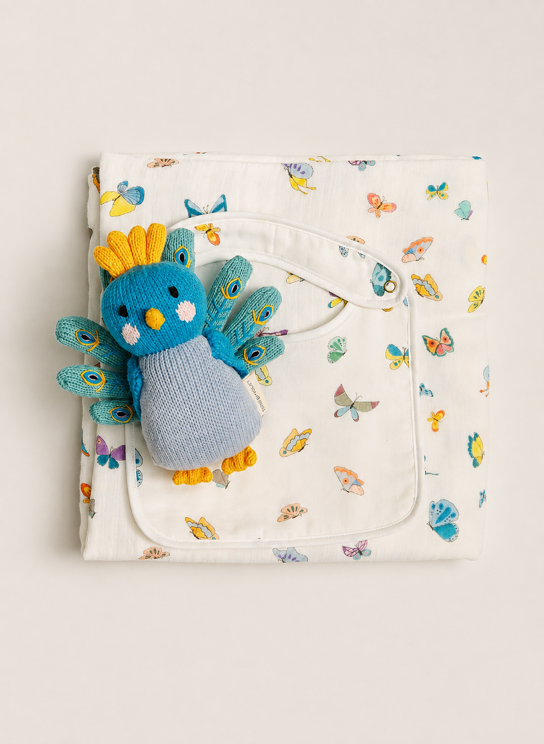 A folded white muslin baby blanket with a colorful butterfly and peacock pattern, featuring a front pocket with a white drawstring and a blue knitted peacock plush toy tucked inside.