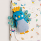 A blue and light blue knitted bird rattle toy with yellow accents, resting on top of a white muslin blanket printed with colorful butterflies.