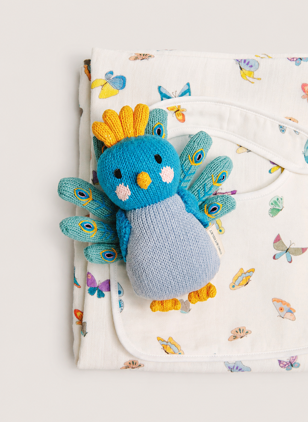 A blue and light blue knitted bird rattle toy with yellow accents, resting on top of a white muslin blanket printed with colorful butterflies.