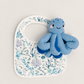 A blue knitted octopus toy resting on a white baby bib with a blue and purple underwater coral print, set against a solid light beige background.