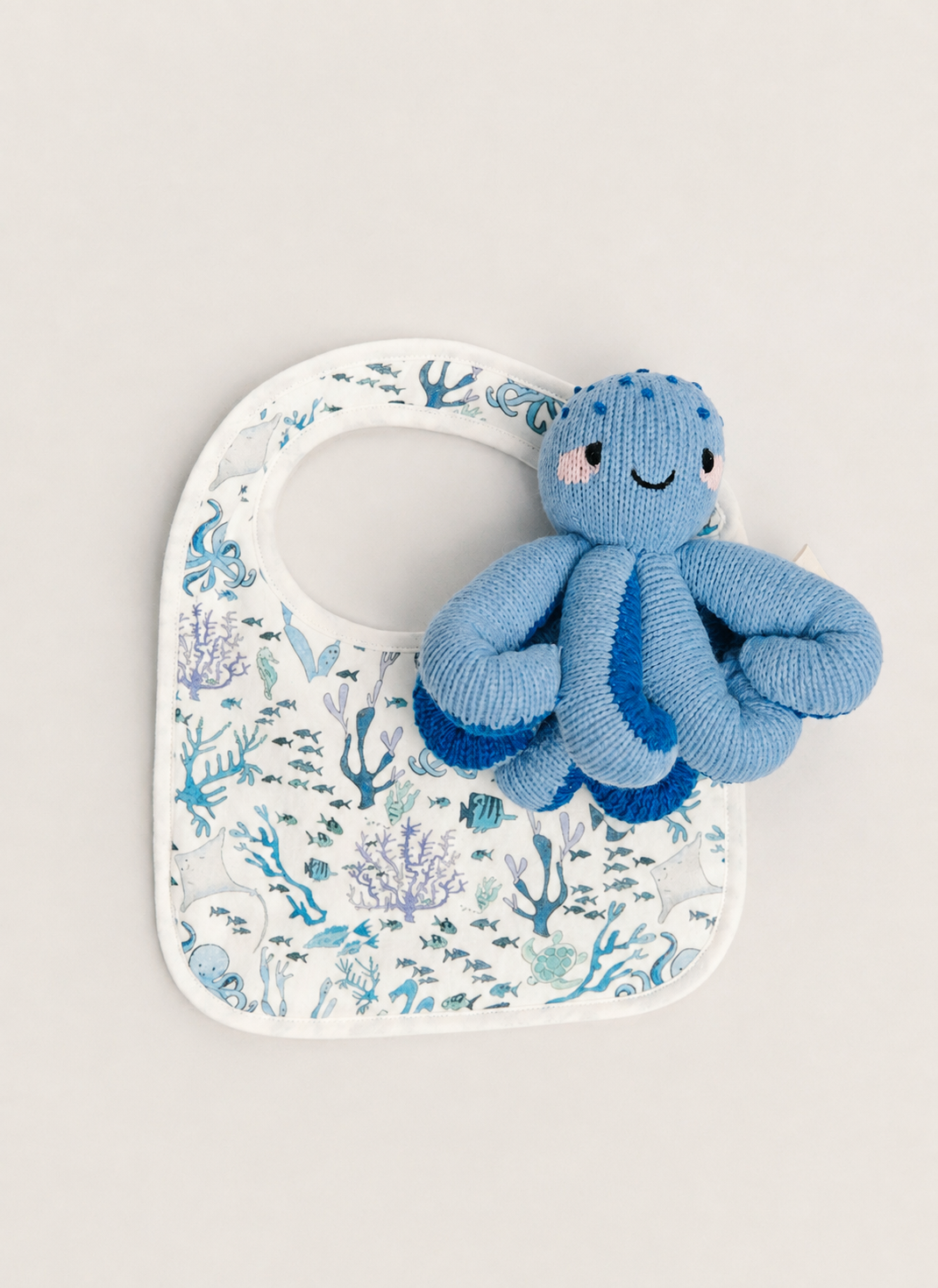 A blue knitted octopus toy resting on a white baby bib with a blue and purple underwater coral print, set against a solid light beige background.