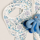 A baby zip-front one-piece outfit in white with an allover blue and teal underwater print, shown with a matching bib and a blue octopus plush toy on a light neutral background. The brand name '10 & VIOLET' is visible near the collar.