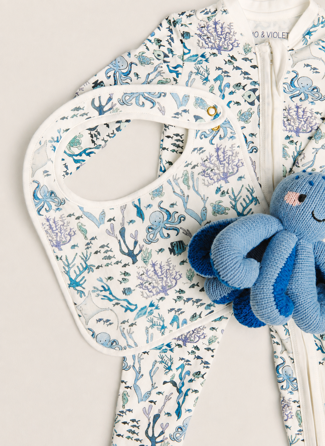 A baby zip-front one-piece outfit in white with an allover blue and teal underwater print, shown with a matching bib and a blue octopus plush toy on a light neutral background. The brand name '10 & VIOLET' is visible near the collar.