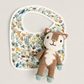 A white baby bib with a colorful woodland animal print, styled with a small knitted plush squirrel toy on a light beige background.