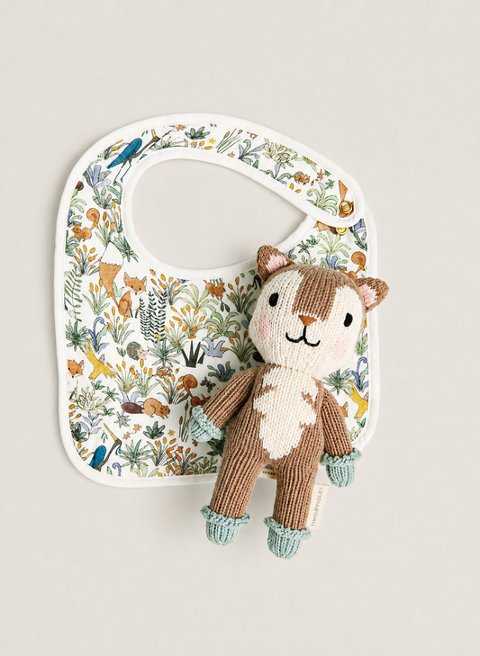 A white baby bib with a colorful woodland animal print, styled with a small knitted plush squirrel toy on a light beige background.