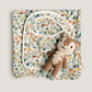 A folded baby blanket or quilt with a dense woodland animal print, styled with a white bib and a small plush teddy bear on a light gray background.