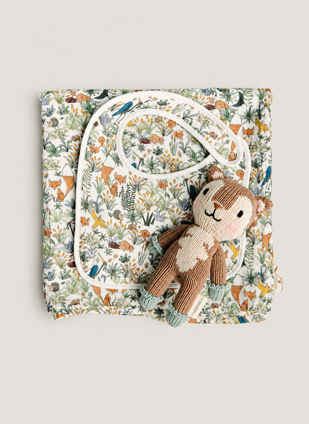 A folded baby blanket or quilt with a dense woodland animal print, styled with a white bib and a small plush teddy bear on a light gray background.