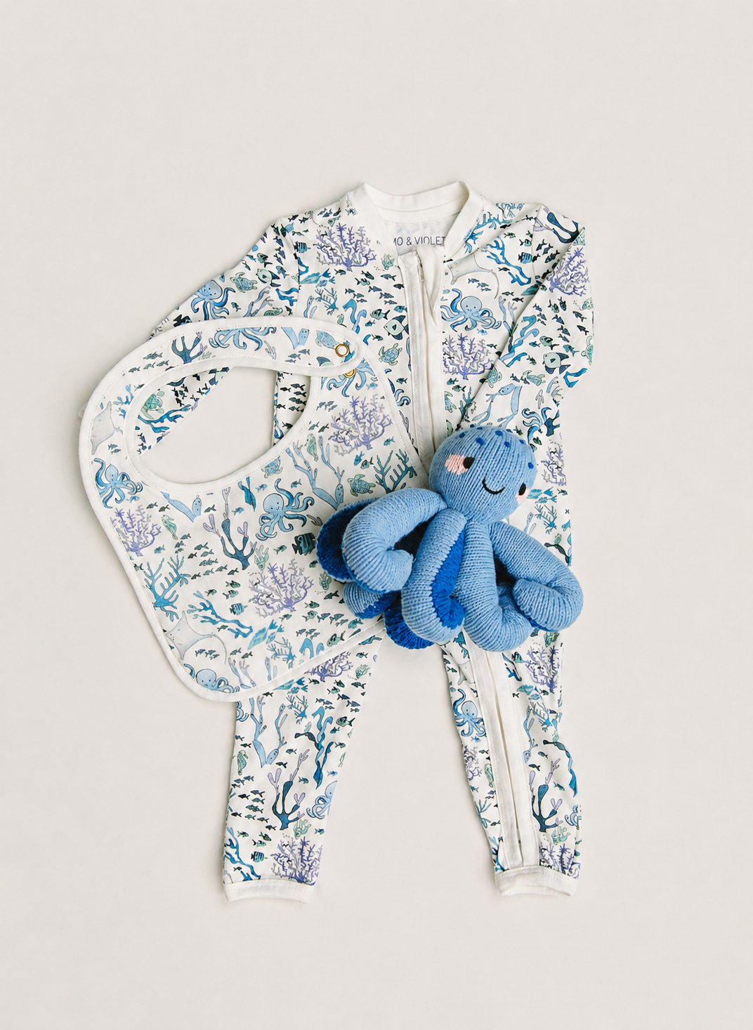 A baby zip-front footed one-piece outfit in an allover blue and purple underwater coral print, shown with a matching bib and a small blue octopus plush toy on a light beige background.