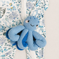A close-up of a baby's footed one-piece sleeper with a white background and a blue and light blue ocean-themed print, featuring a smiling blue knitted octopus toy resting on the zipper.