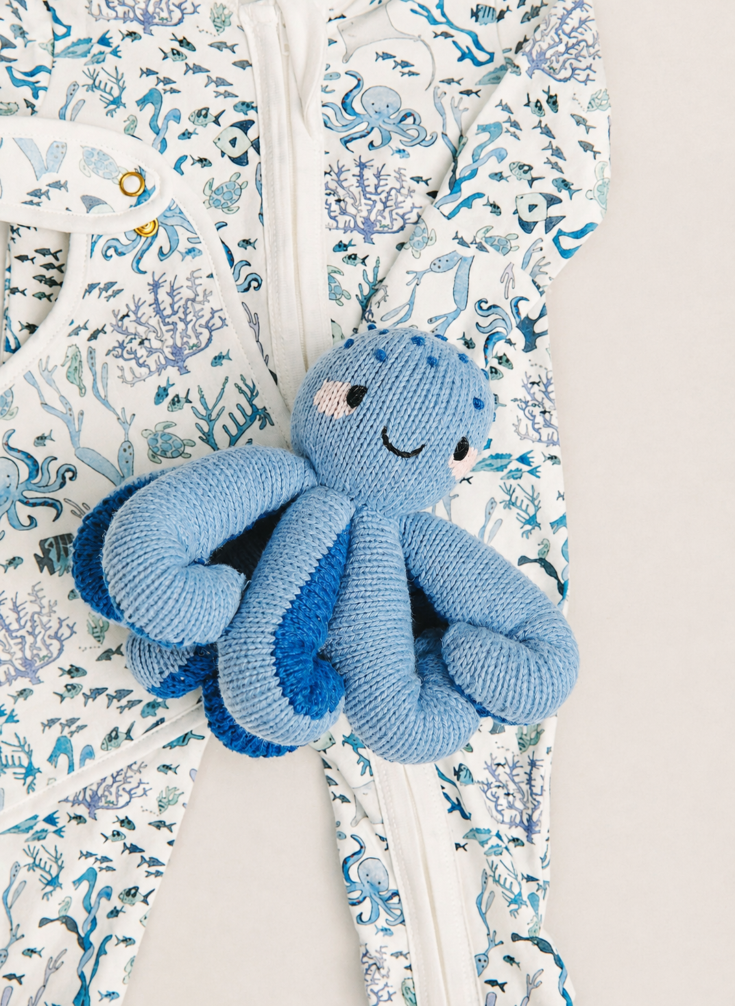 A close-up of a baby's footed one-piece sleeper with a white background and a blue and light blue ocean-themed print, featuring a smiling blue knitted octopus toy resting on the zipper.