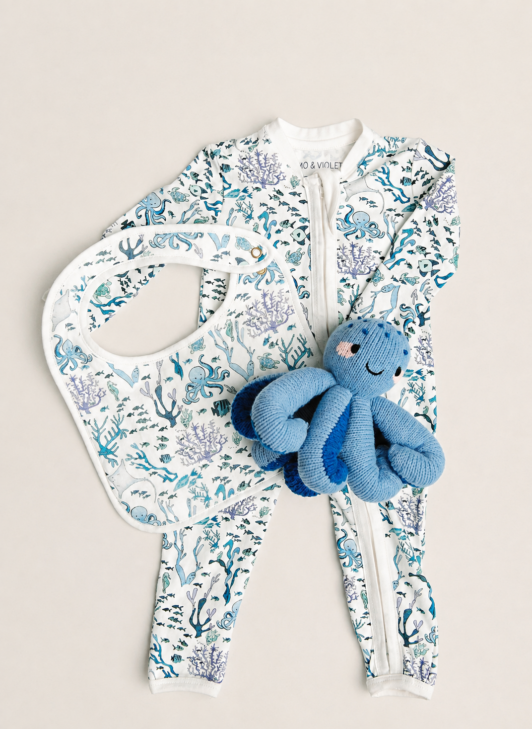 A baby outfit set featuring a white one-piece jumpsuit with a blue and purple sea-creature print, a matching bib, and a blue octopus plush toy, arranged on a light beige background.