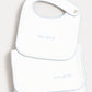 A white baby bib and matching folded cloth napkin set, both embroidered with the word 'amore' in light blue script and finished with a light blue scalloped edge.