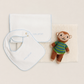 A baby gift set featuring a white muslin bib with light blue scalloped edges and embroidered 'smile' in blue, a white muslin square cloth with light blue scalloped edges, a textured cream baby blanket, and a small brown plush monkey toy wearing a green and blue striped shirt, all arranged on a warm beige background.