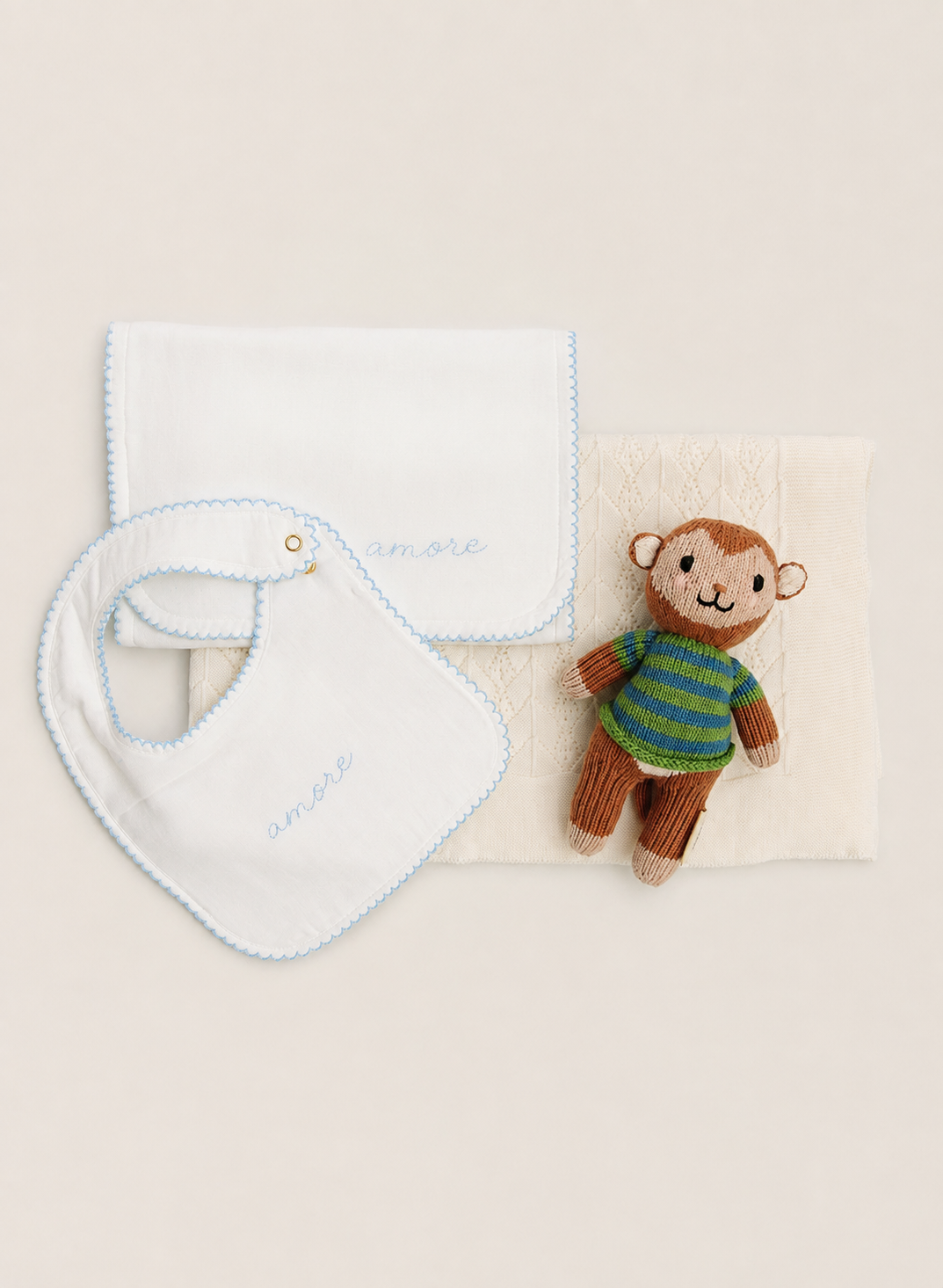 A baby gift set featuring a white muslin bib with light blue scalloped edges and embroidered 'smile' in blue, a white muslin square cloth with light blue scalloped edges, a textured cream baby blanket, and a small brown plush monkey toy wearing a green and blue striped shirt, all arranged on a warm beige background.