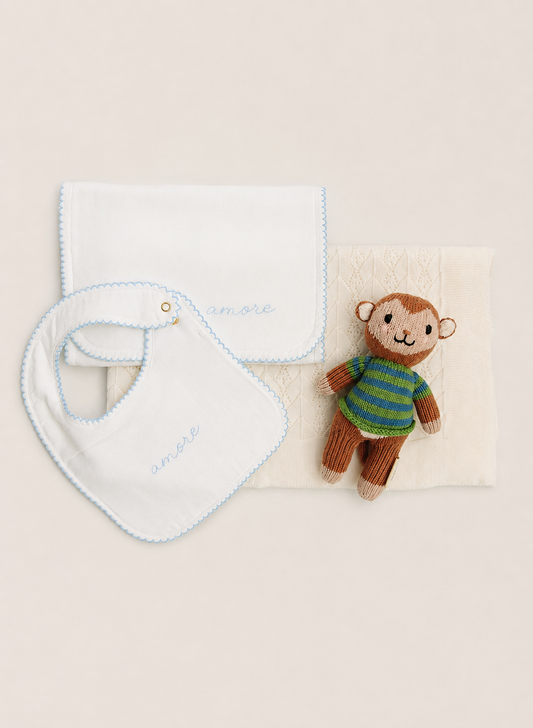A baby gift set featuring a white muslin bib with light blue scalloped edges and embroidered 'smile' in blue, a white muslin square cloth with light blue scalloped edges, a textured cream baby blanket, and a small brown plush monkey toy wearing a green and blue striped shirt, all arranged on a warm beige background.
