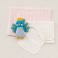 A baby gift set featuring a light pink knitted blanket, a white cotton bib with a snap closure, and a small blue and yellow peacock plush toy, arranged neatly on a light beige background.