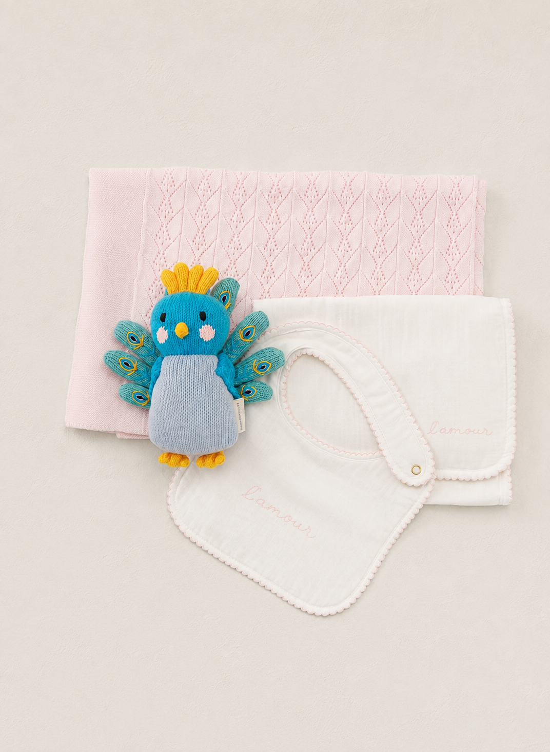 A baby gift set featuring a light pink knitted blanket, a white cotton bib with a snap closure, and a small blue and yellow peacock plush toy, arranged neatly on a light beige background.