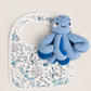 A white baby bib with an ocean-themed print of blue and purple fish, coral, and sea life, styled with a blue knitted octopus toy on top, set against a solid light beige background.