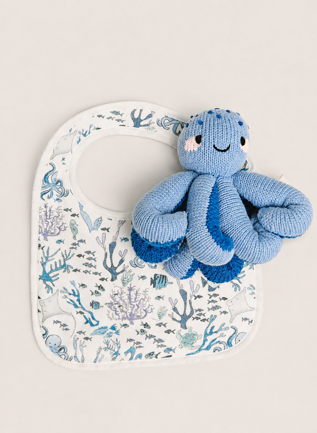 A white baby bib with an ocean-themed print of blue and purple fish, coral, and sea life, styled with a blue knitted octopus toy on top, set against a solid light beige background.