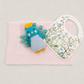 A baby gift set featuring a pink textured blanket, a white patterned bib with colorful bird illustrations, and a blue and yellow plush peacock toy, arranged on a neutral light gray background.