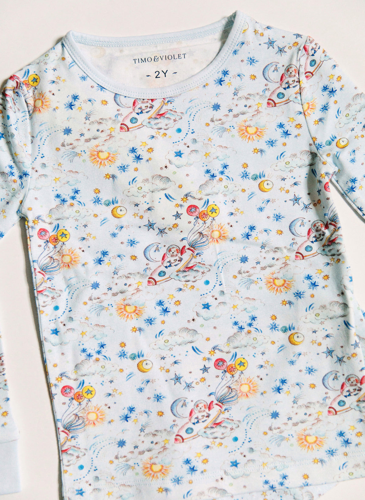 A children's long-sleeve top by Timo & Violet, featuring a colorful space-themed print with rockets, planets, stars, and suns on a pale blue background.