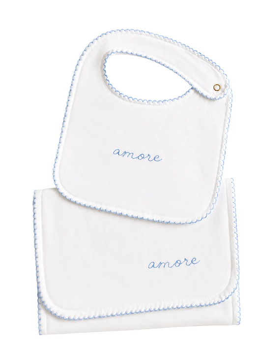 A white baby bib and matching footie cloth set with light blue scalloped edges and the word 'amore' embroidered in blue on each piece, shown on a warm off-white background.