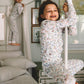 A child wearing a floral print long-sleeve pajama set climbs on a white pole in a bedroom, with another child in matching pajamas visible in the background near a bed.
