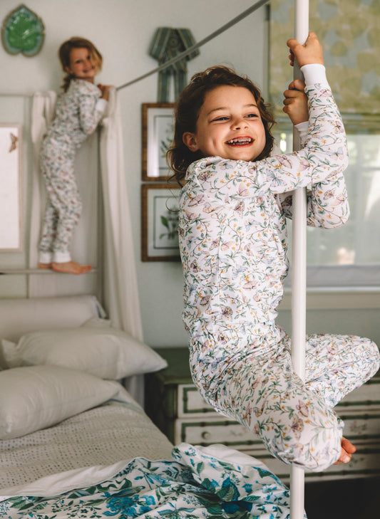 A child wearing a floral print long-sleeve pajama set climbs on a white pole in a bedroom, with another child in matching pajamas visible in the background near a bed.