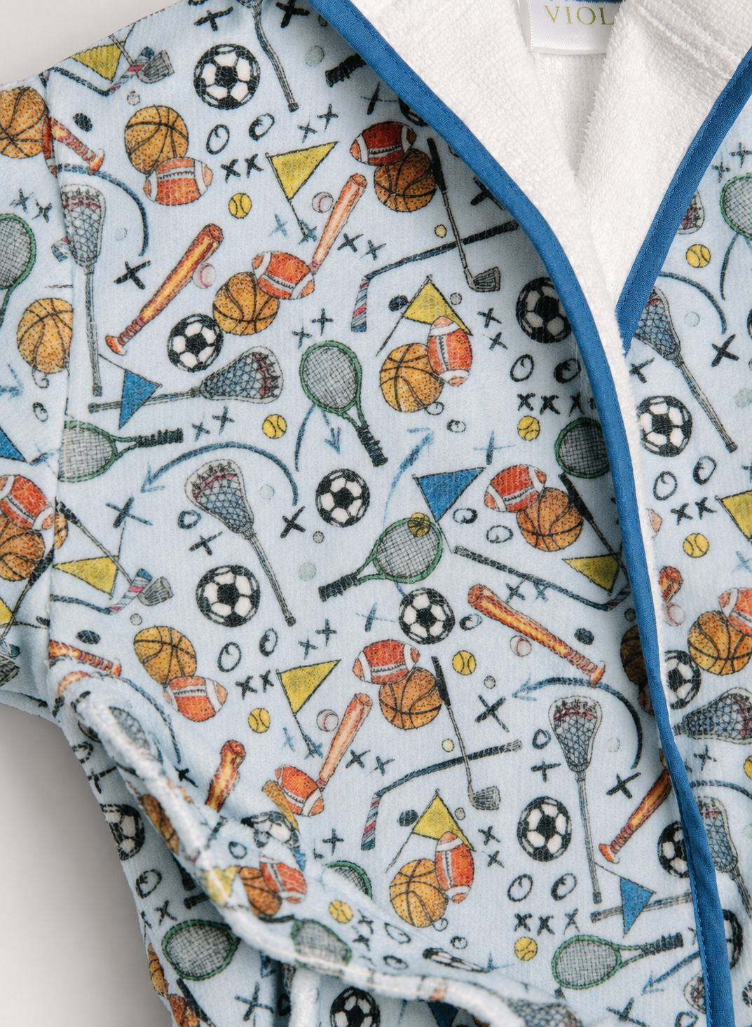 A close-up of a light blue fabric garment with a multicolor sports-themed print featuring basketballs, soccer balls, lacrosse sticks, rackets, and balls, finished with a white plush lining and blue piping.